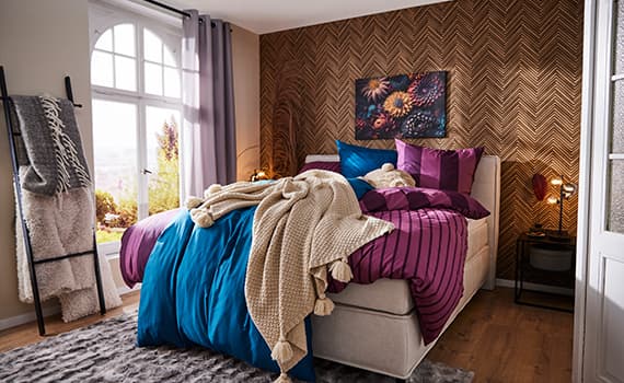 Gemütliches Schlafzimmer mit Doppelbett, gestreifter Bettwäsche, blauen und violetten Kissen, Decke und großem Fenster.