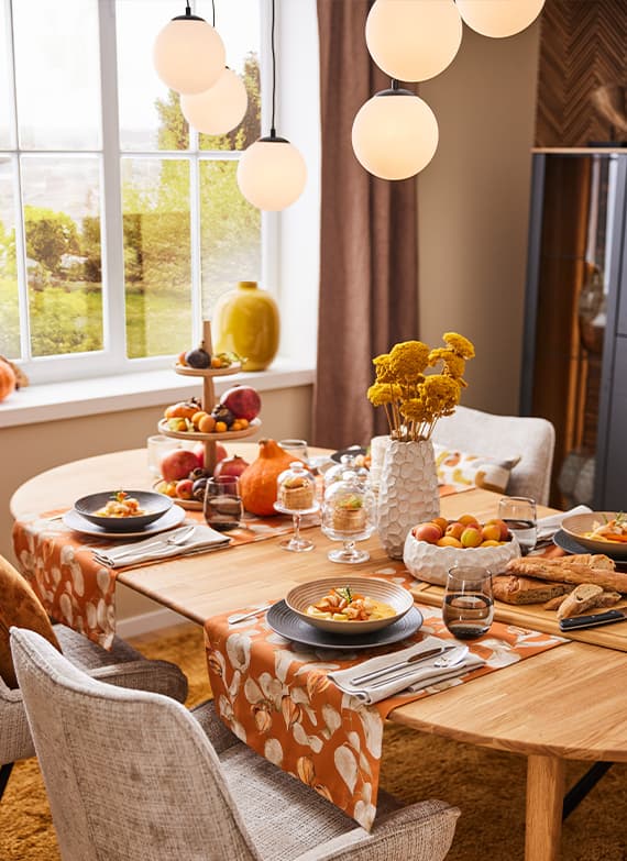 Festlich gedeckter Esstisch mit herbstlichen Deko-Elementen, Tellern mit Essen, Obst, Brot und Blumen in Vasen vor einem Fenster.