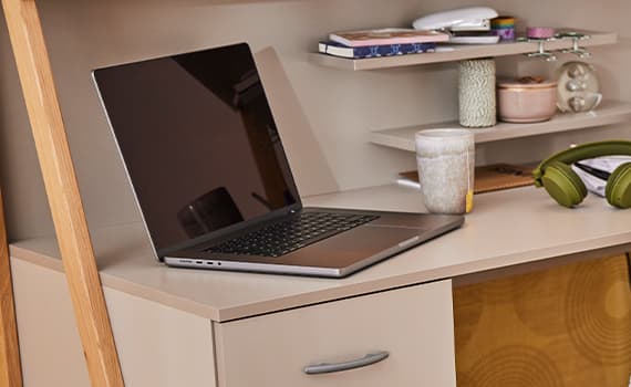 Offener Laptop auf einem Schreibtisch neben einer Tasse, grünen Kopfhörern und Regal mit Büchern und Dekoration