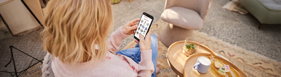 Frau sitzt auf Sofa und schaut auf ihr Smartphone, auf Couchtisch stehen Kaffee und Snacks