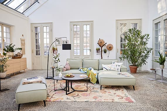 Helles Wohnzimmer mit großer Eckcouch, Hocker, Couchtischen, Teppich und vielen grünen Pflanzen vor hohen Fenstern