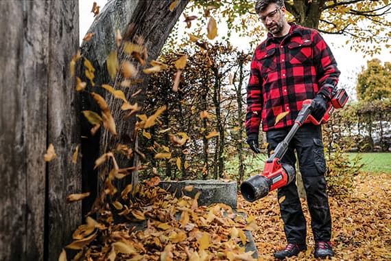 Mann im karierten Hemd bläst mit Akku-Laubbläser herabgefallene Blätter unter einem Baum im herbstlichen Garten weg