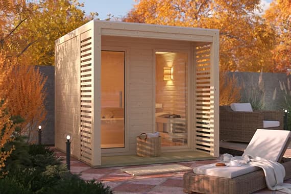 Gartensauna mit Glasfront auf Terrasse, davor Liegestuhl und Korb, umgeben von herbstlichen Bäumen und Sitzgruppe