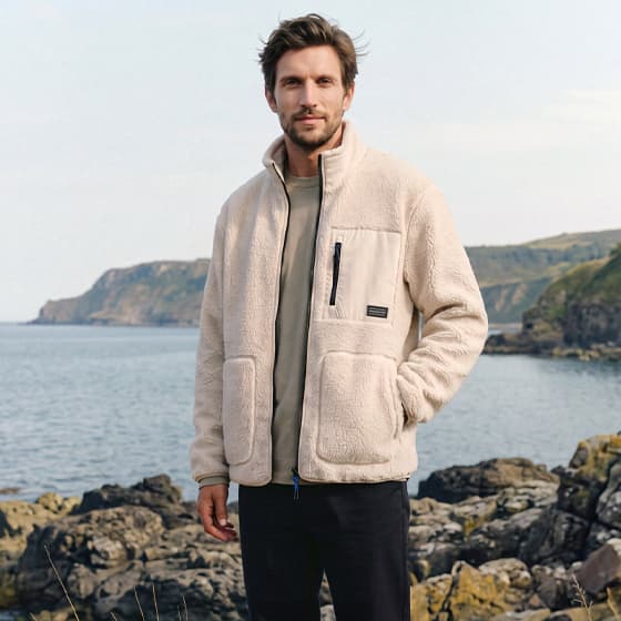 Mann in heller Jacke steht mit Händen in den Taschen auf Felsen am Meer, Küste und Hügel im Hintergrund