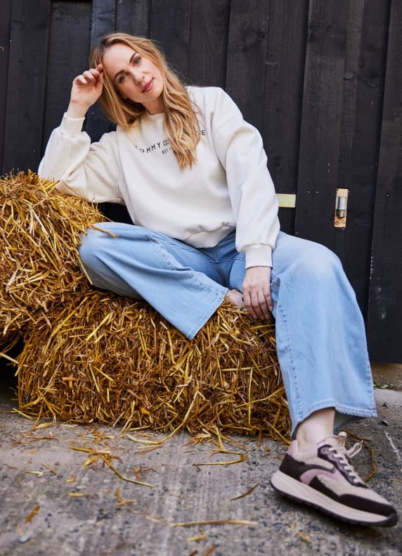Frau im bedruckten Sweatshirt und weitem Jeans sitzt entspannt auf Strohballen vor dunkler Holzwand