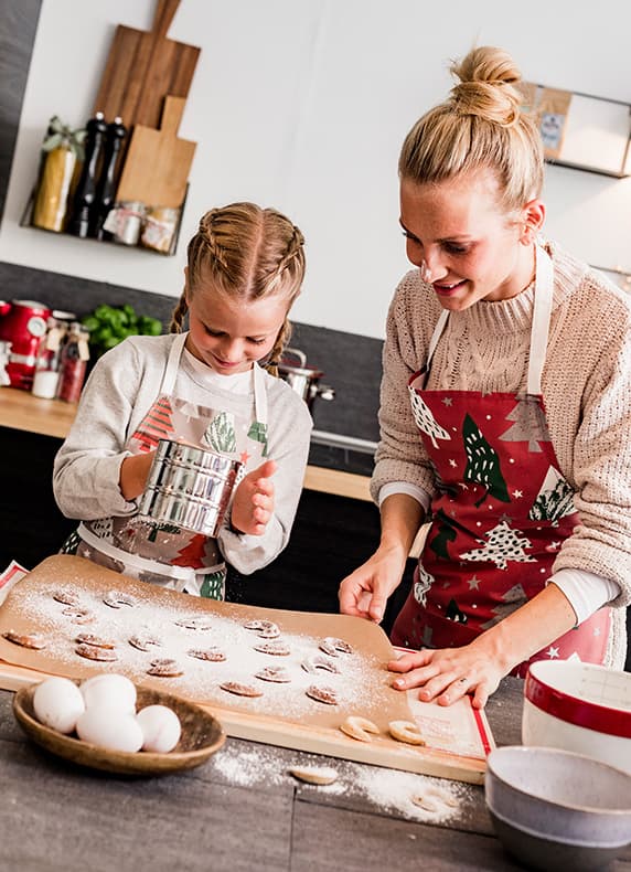 Frau und Mädchen mit Weihnachtschürzen bestäuben Plätzchen auf einem Backblech in moderner Küche mit Eiern im Vordergrund