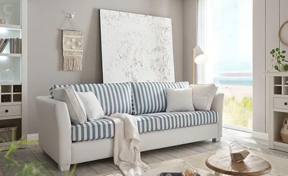 Wohnzimmer mit weißer Couch und blau-weiß gestreiften Kissen, hellem Teppich, Wanddeko, großer Fensterfront und Stehlampe