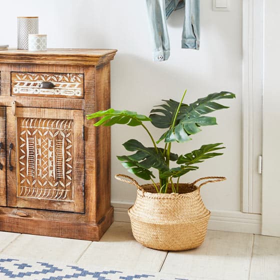 Geflochtener Korb mit grüner Pflanze neben Holzschrank mit geschnitzten Mustern, darüber hängende Jeansjacke