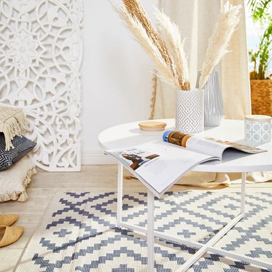 Wohnzimmer mit rundem Tisch, darauf Magazin, Vase mit Pampasgras, Tasse und Schale, dahinter dekoratives weißes Wandornament.