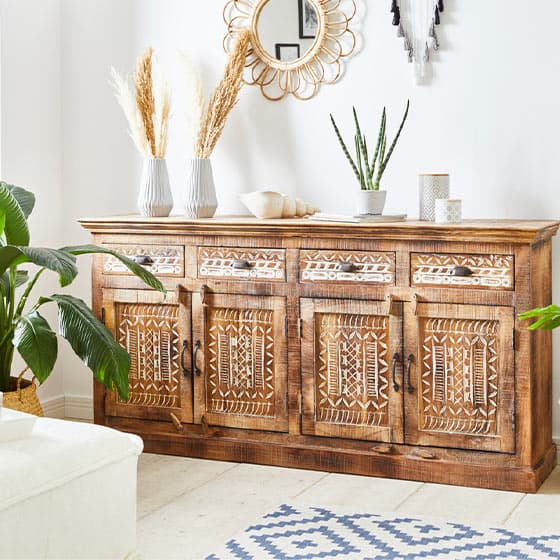 Rustikale Holzkommode mit kunstvollen Mustern, dekoriert mit Vasen, Pflanzen und einem Spiegel darüber in einem hellen Raum.