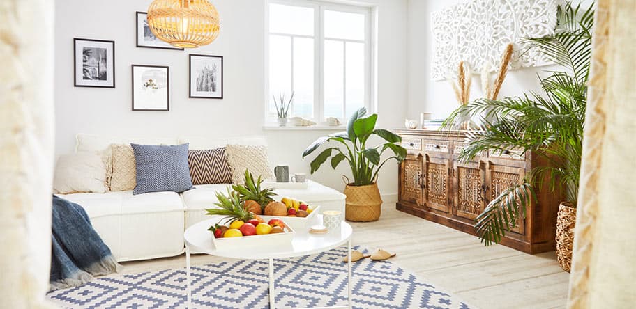 Helles Wohnzimmer mit Sofa, Kissen, Pflanzen, Holzkommode und rundem Tisch voller Früchte auf gemustertem Teppich.
