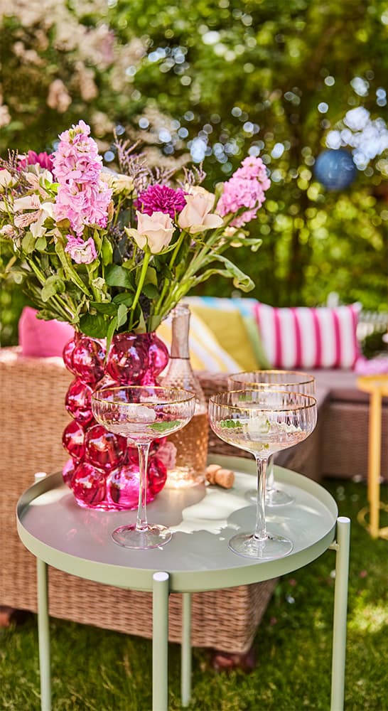 Blumenstrauß in rosa Vase, zwei Champagnergläser und eine Flasche auf Tisch, gemütliche Sitzgruppe im Garten im Hintergrund