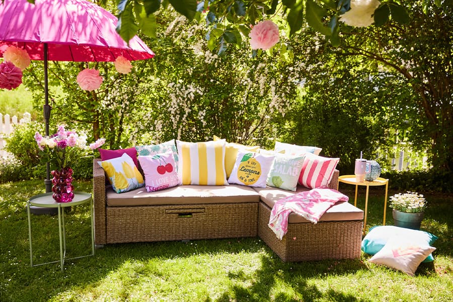 Gemütliche Gartenlounge mit bunten Kissen unter pinkem Sonnenschirm, umgeben von grünen Bäumen und Blumen im Sonnenlicht