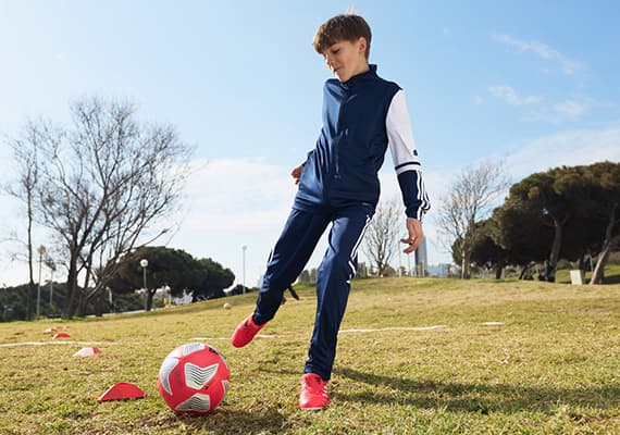 Junge in blau-weißem Sportanzug spielt Fußball auf einer Wiese, roter Fußball und Hütchen auf dem Boden, sonniger Tag.