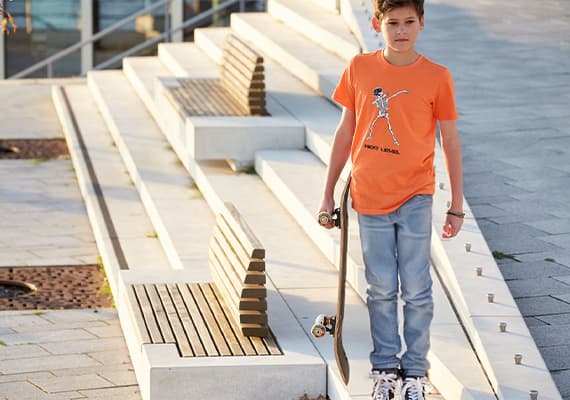 Junge in orangefarbenem T-Shirt und blauen Jeans hält ein Skateboard neben einer Treppe mit Holzsitzen.