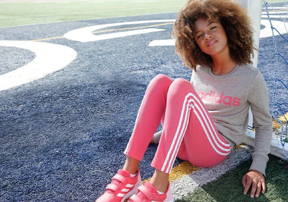 Mädchen sitzt auf Sportplatz, trägt graues Adidas-Sweatshirt, pinke Adidas-Hose mit weißen Streifen und rosa Turnschuhe.