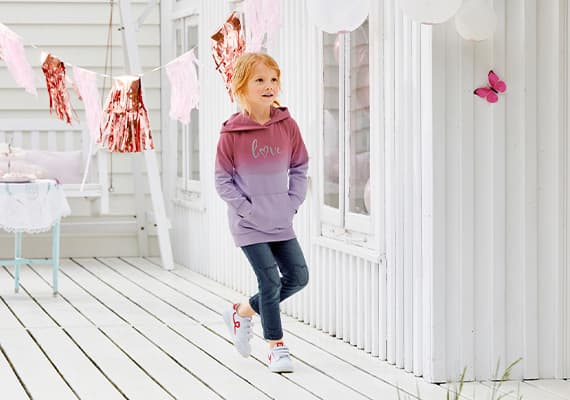 Kind in lila-rosa Kapuzenpullover mit "love"-Schriftzug, Jeans und weißen Sneakern steht auf weißer Holzterrasse vor Haus.