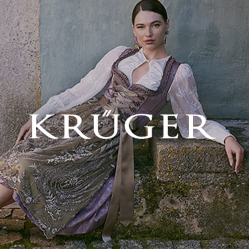 KRÜGER – Frau in traditioneller Tracht lehnt lässig an einer Steinmauer