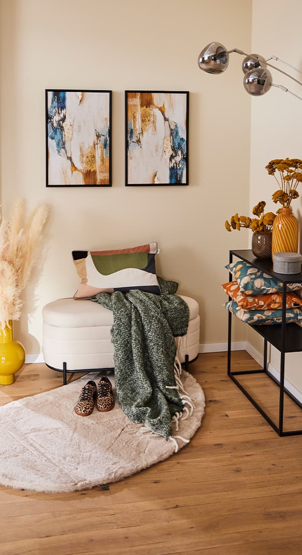 Wohnzimmer mit beiger Bank, grüner Decke, gemusterten Kissen und Leoparden-Schuhen auf Teppich. Zwei abstrakte Bilder an der Wand.