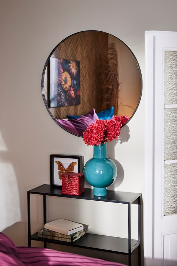 Runder Spiegel über schwarzem Regal mit rotem Korb, Buch, Schmetterlingsbild und Vase mit roten Blumen