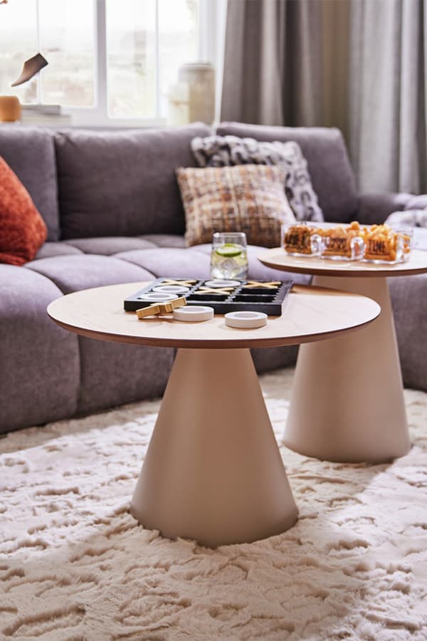 Zwei runde Beistelltische mit Spiel und Snacks darauf, im Hintergrund graues Sofa mit bunten Kissen und Fenster mit Vorhängen.