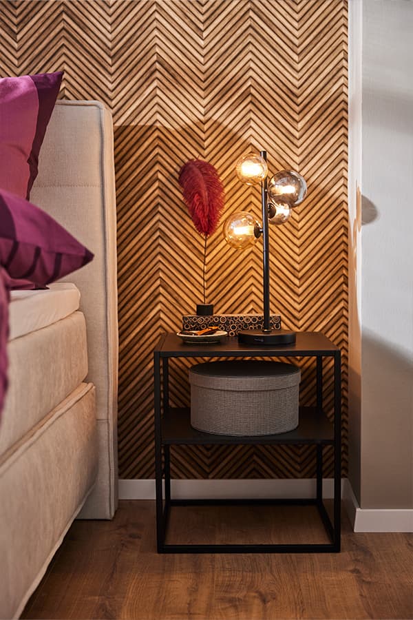 Nachttisch mit moderner Lampe und Deko vor Wand mit Fischgrätmuster neben Bett mit violettem Kissen