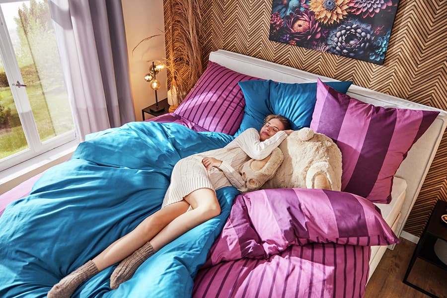 Person in cremefarbenem Pullover und Socken liegt auf einem Bett mit blau-lila Bettwäsche, Kuscheltier im Arm, neben Fenster und Lampe.