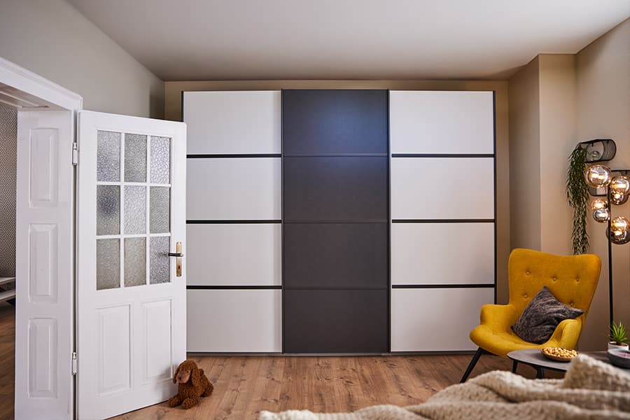 Modernes Schlafzimmer mit großem Kleiderschrank, gelbem Sessel, Kissen, rundem Tisch, Dekolampe und Hundespielzeug auf dem Boden.