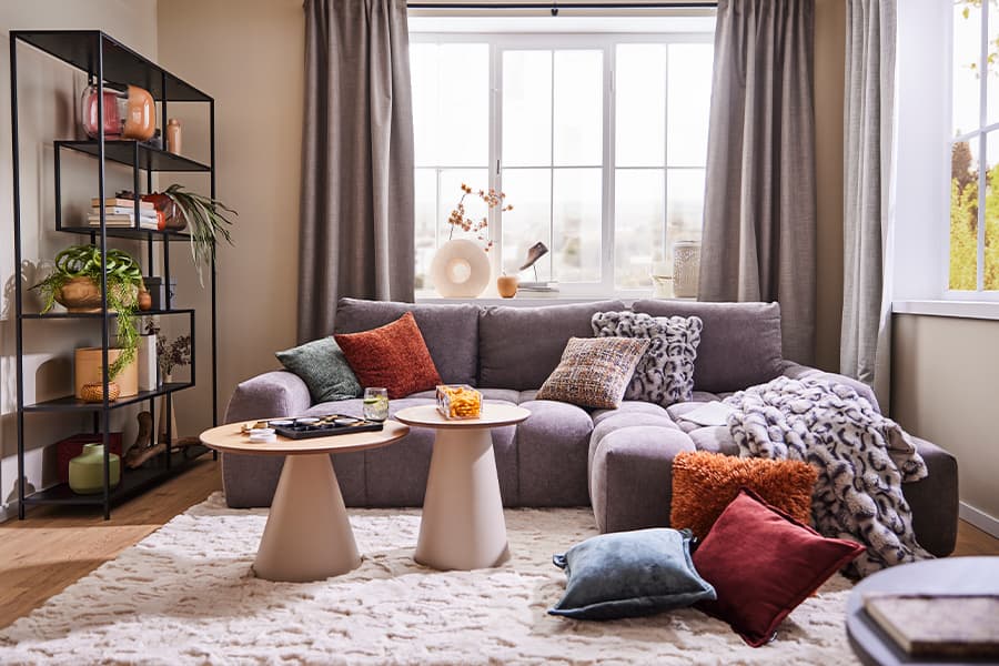 Gemütliches Wohnzimmer mit grauem Sofa, bunten Kissen und zwei runden Beistelltischen vor einem großen Fenster mit Vorhängen