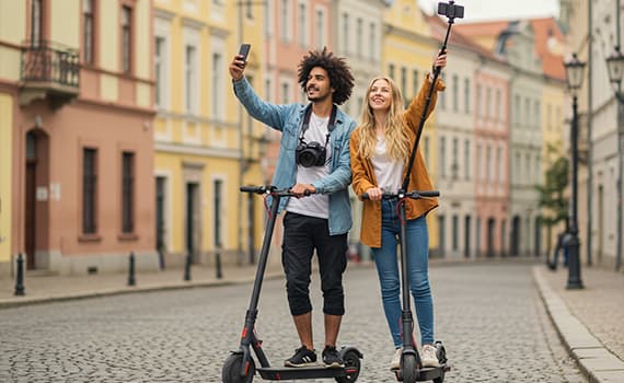 Zwei Personen auf E-Scootern machen Selfies in einer malerischen Altstadt mit bunten Gebäuden und gepflasterten Straßen.