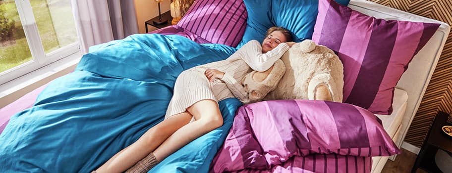 Person in cremefarbenem Pulloverkleid liegt auf Bett mit blau-lila Bettwäsche neben einem Plüschbären.