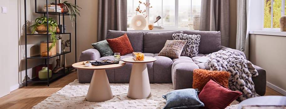 Gemütliches Wohnzimmer mit grauem Sofa, bunten Kissen und zwei runden Couchtischen, Fenster im Hintergrund, Regal links