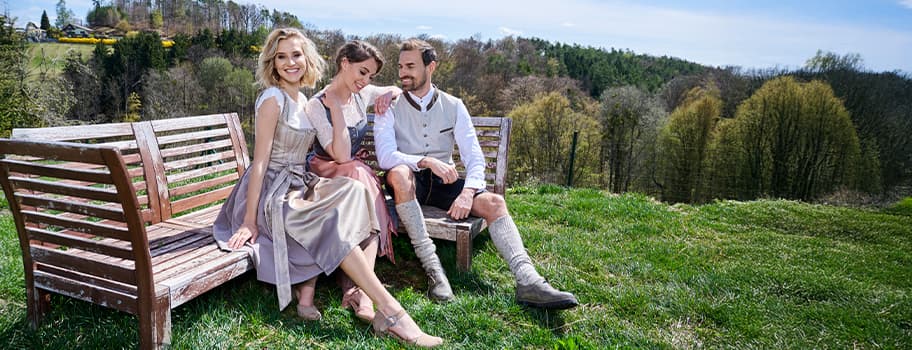 Zwei Frauen und ein Mann in Tracht sitzen auf Holzbänken im Grünen vor einer Waldlandschaft.