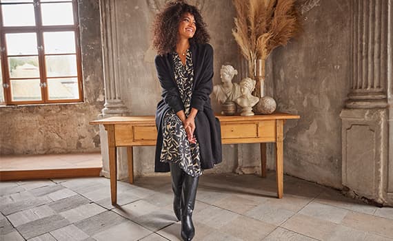 Curvy Frau mit lockigem Haar sitzt auf Tisch in rustikalem Raum, trägt gemustertes Kleid, schwarzen Cardigan und Stiefel.