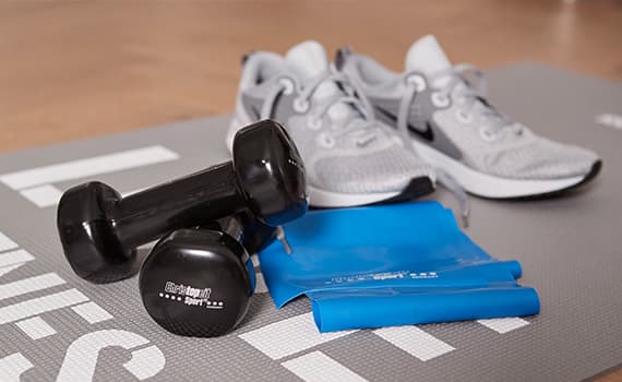 Zwei schwarze Hanteln, blaues Widerstandsband und graue Sportschuhe auf grauer Fitnessmatte