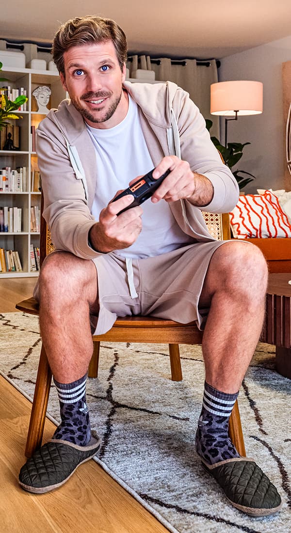 Mann sitzt in gemütlichem Wohnzimmer und hält einen TV-Controller in der Hand, trägt bequeme Kleidung und Hausschuhe