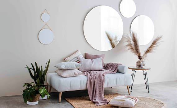 Gemütliches Wohnzimmer mit grauem Sofa, rosa Decke, Kissen, Pflanzen und runden Spiegeln an der Wand.