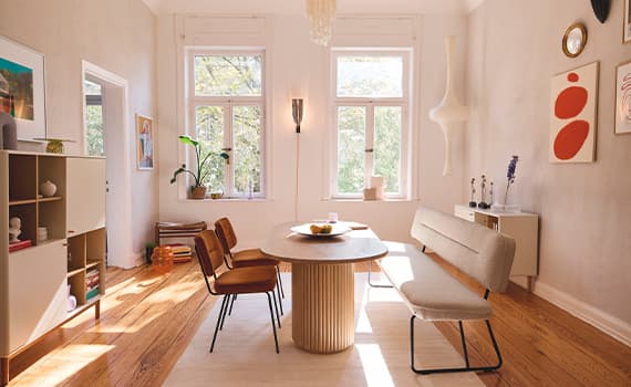 Esszimmer mit runden Tisch, braunen Stühlen und grauer Bank, Holzregal links, Pflanzen und Fenster mit Blick ins Grüne