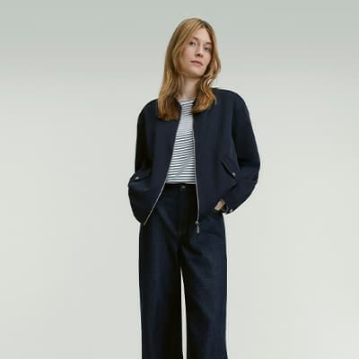 Frau trägt gestreiftes Shirt, dunkelblaue Jacke und weite dunkelblaue Hose, Hände in Jackentaschen, Gesicht unscharf