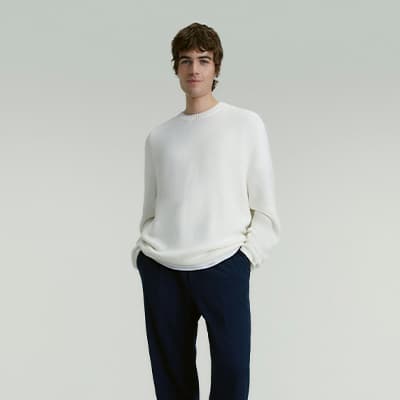 Person in weißem Pullover und dunkler Hose steht mit Händen in den Taschen vor neutralem Hintergrund