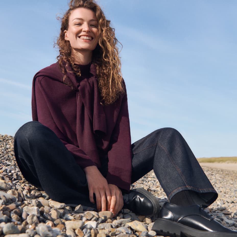 Frau in dunkelrotem Pullover und dunkler Jeans sitzt auf Kieselsteinen am Strand, blauer Himmel im Hintergrund