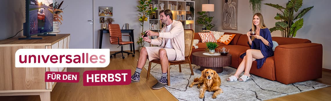 Mann spielt Videospiel im Wohnzimmer, Frau sitzt auf rotem Sofa und isst, Hund liegt auf Teppich, Möbel und Pflanzen im Hintergrund