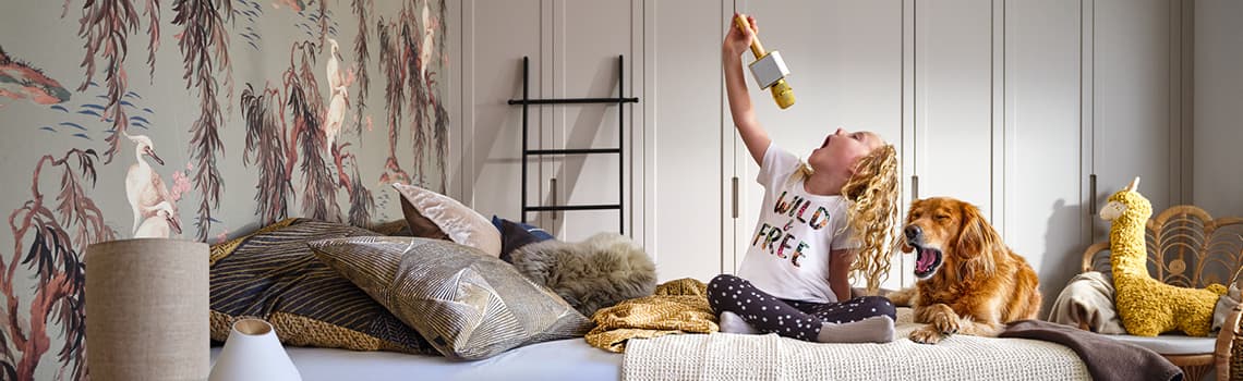 Kind in "Wild Free" T-Shirt hält Spielzeugmikrofon hoch, sitzt auf Bett mit Hund und Kissen, gemusterte Tapete im Hintergrund