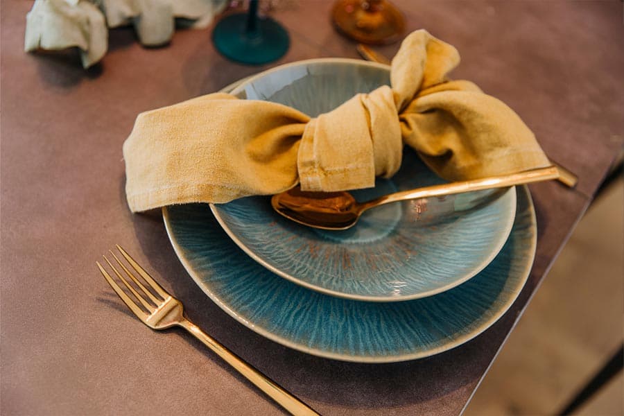 Elegant gedeckter Tisch mit goldfarbenem Besteck, blauem Teller, gelber Stoffserviette und braunem Löffel auf Keramikteller.