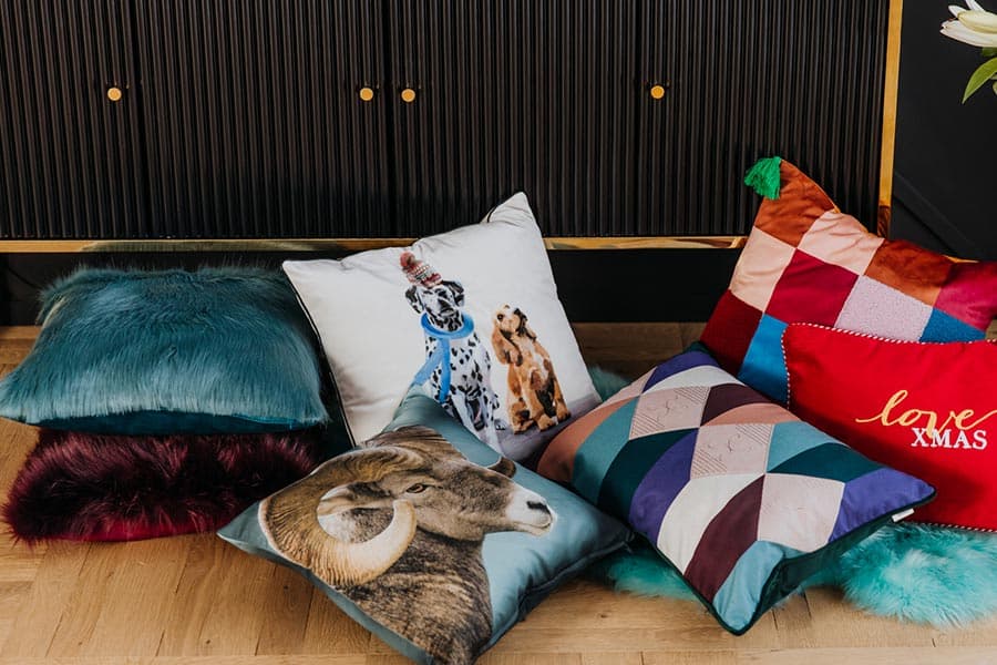 love XMAS – verschiedene dekorative Kissen, darunter Tiere und geometrische Muster, auf dem Boden vor einem dunklen Möbelstück