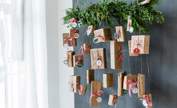DIY Adventskalender aus kleinen braunen Geschenken, aufgehängt an einem grünen Zweig mit Nummern und Schleifen verziert