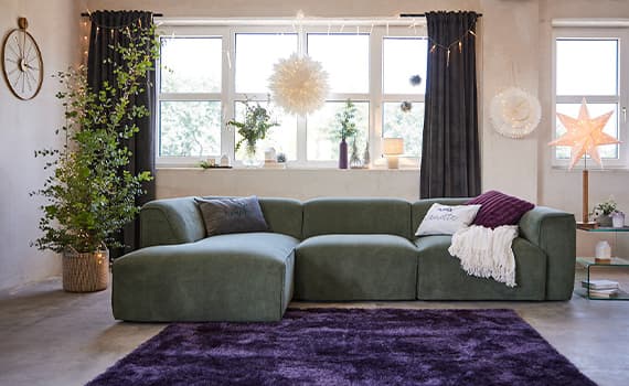 Grünes Ecksofa mit Kissen und Decke, lila Teppich, Fenster mit Pflanzen und Dekoration, große Stern- und Pompon-Lichter