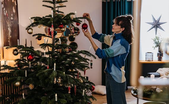 Person schmückt Weihnachtsbaum mit roten Kugeln und Kerzen in gemütlichem Wohnzimmer mit weihnachtlicher Dekoration