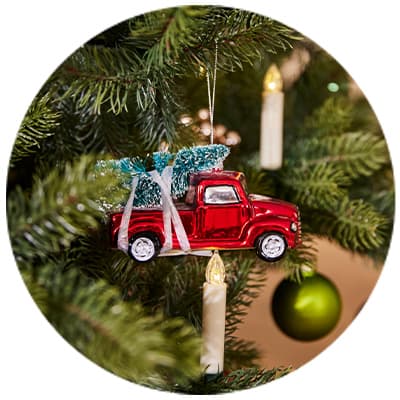Rot glänzender Spielzeug-Truck mit Tannenbaum auf Ladefläche hängt als Christbaumkugel an Weihnachtsbaum neben Kerzen und grüner Kugel