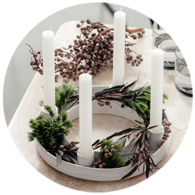 Weißer Adventskranz mit vier weißen Kerzen und grünen Zweigen auf einem Tisch, daneben Gläser und Dekoration.